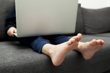 Çocuk kanepede oturmuş dizüstü bilgisayarını tutuyor ve izliyor. 