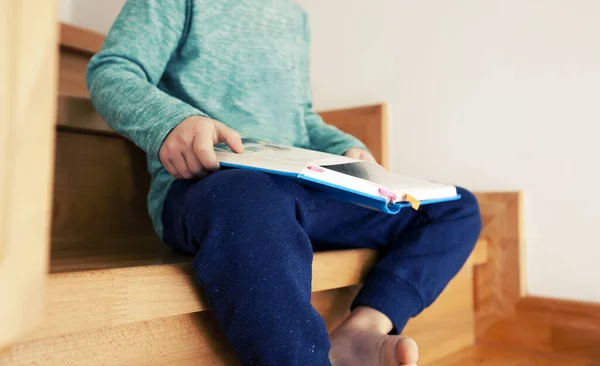 The child is reading the paper book sitting on the wooden stairs in the house