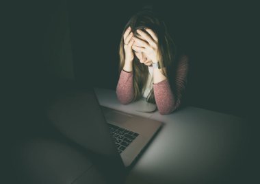Çok yorgun ve bitkin bir kadın, gece geç saatte dizüstü bilgisayarın başında elleriyle başını tutuyor. İşkolik, aşırı çalışma ve endişe kavramı