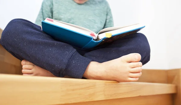 The child is reading the paper book sitting on the wooden stairs in the house