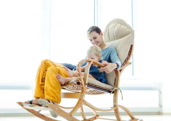 Mother and daughter sitting in a rocking chair. Indoor photo on the background of a large bright window.