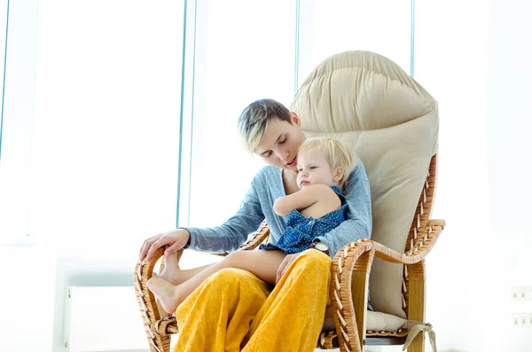 Mother and daughter sitting in a rocking chair. Indoor photo on the background of a large bright window.