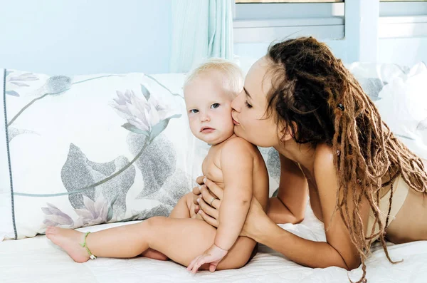 Moeder en dochter liggen op het bed. Vrouw en kind in een lichte kamer. Meisje met dreadlocks. — Stockfoto