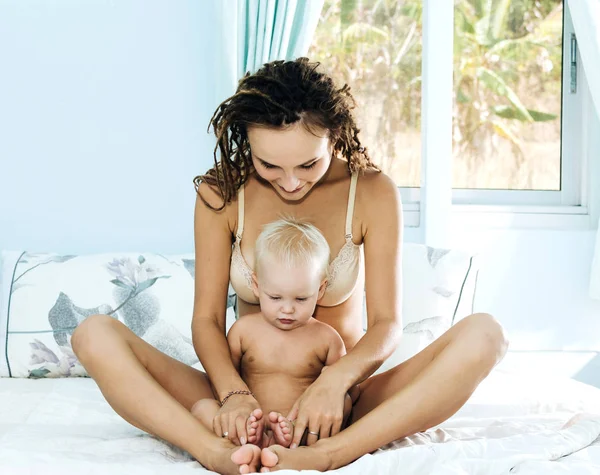 Moeder en dochter liggen op het bed. Vrouw en kind in een lichte kamer. Meisje met dreadlocks met gekruiste benen zit en het houden van een baby. — Stockfoto