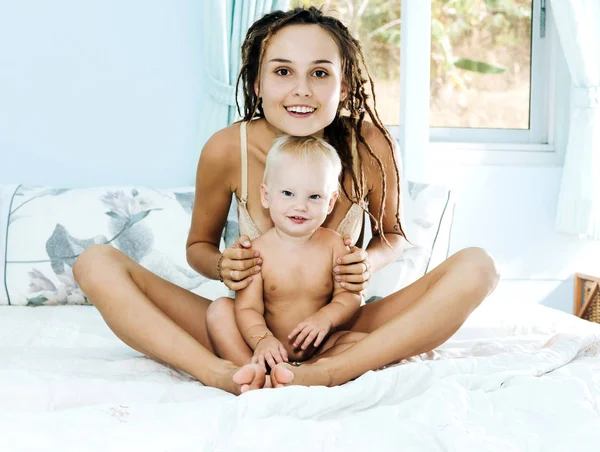 Мама і дочка лежати на ліжку. Жінка і дитина у світлій кімнаті. Дівчина з дреди сидить схрестивши ноги і тримає дитину. — стокове фото
