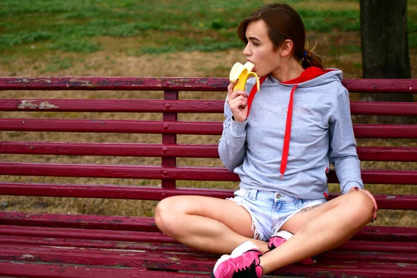 Ein junges süßes Mädchen ist sehr verführerisch, wenn es eine Banane isst. eine Sportlerin beobachtet ihr Essen. — Stockfoto