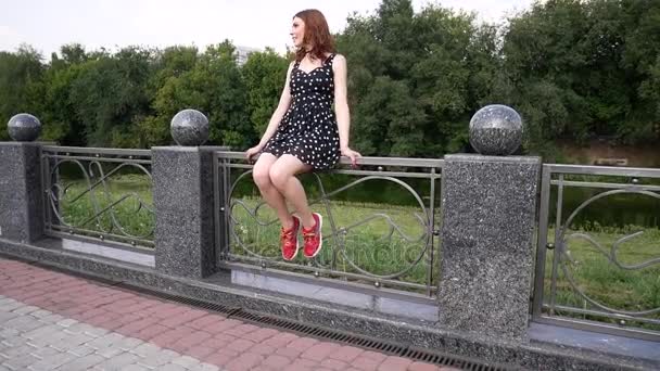 Flickan sitter på räcket och skakar benen. Vacker flicka i en sexig klänning, sorglösa promenader på gatan. — Stockvideo