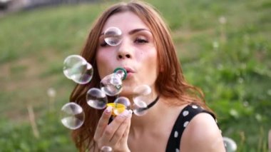 Sabun köpüğü üfleme kız. Genç güzel kadın parkta çim üzerinde oturur ve kabarcıkları ile oynuyor. Slow motion video.