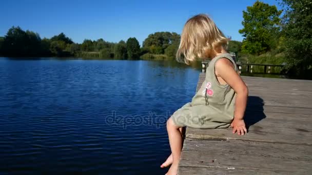 La ragazza è seduta sul molo e chiacchiera con i piedi. Bambino su un ponte di legno vicino all'acqua — Video Stock