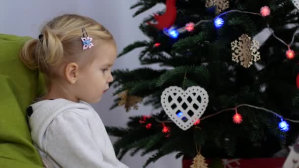 El niño está sentado cerca del árbol de Navidad. El chico está esperando el regalo. — Vídeos de Stock