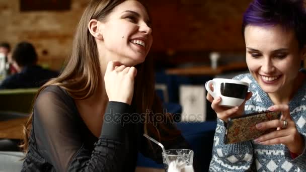Deux amis dans un café regardent la vidéo sur le smartphone et rient. Les femmes boivent du café dans le café — Video