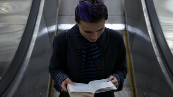 A menina sobe a escada rolante no metrô e lê o livro — Vídeo de Stock