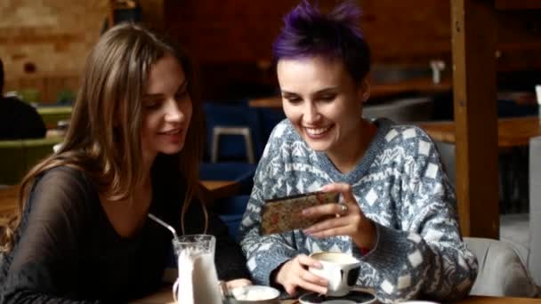 Two friends in a cafe watch video on the smartphone and laugh. Women drink coffee in the coffee house — Stock Video