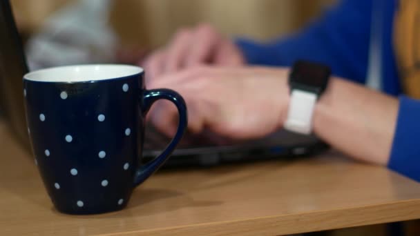 Un pigiste travaille sur un ordinateur portable. Près d'une tasse avec une boisson chaude. Un homme boit du thé et enlève une montre intelligente . — Video