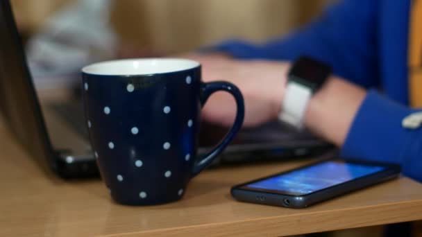 Freelancer está trabajando en un portátil. Cerca de una taza con una bebida caliente. Un hombre bebe té y se quita un reloj inteligente . — Vídeos de Stock