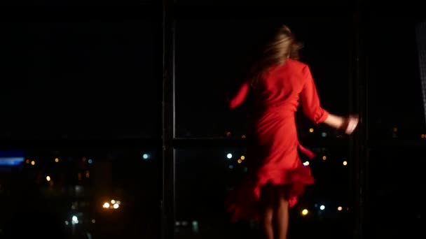 Una chica en un vestido elegante rojo está bailando contra el telón de fondo de una gran ventana con una ciudad de la noche. Sola en casa y enamorada — Vídeos de Stock