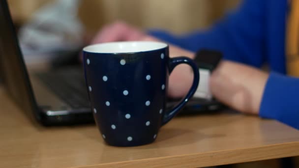 Freelancer está trabajando en un portátil. Cerca de una taza con una bebida caliente. Un hombre bebe té y se quita un reloj inteligente . — Vídeo de stock