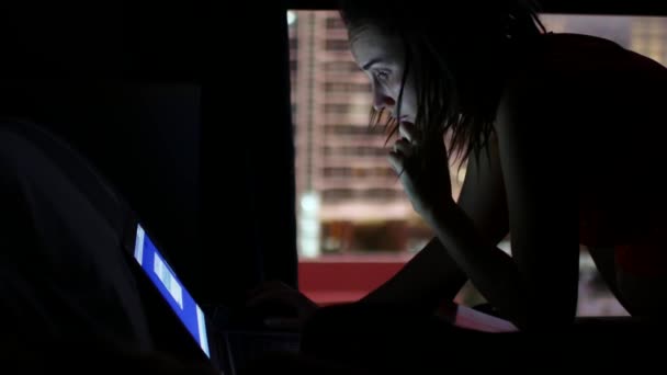 Een vrouw die in het donker aan een laptop werkt. Silhouet op een achtergrond van een raam met uitzicht op de nachtstad — Stockvideo