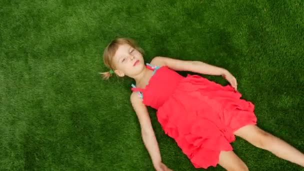 La petite fille est allongée sur de l'herbe verte, vue du dessus. L'enfant fait un ange comme dans la neige — Video