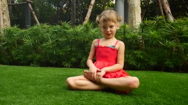 Baby girl is sitting on the grass in lotus position. Girl meditates and sings OM — Stock Video