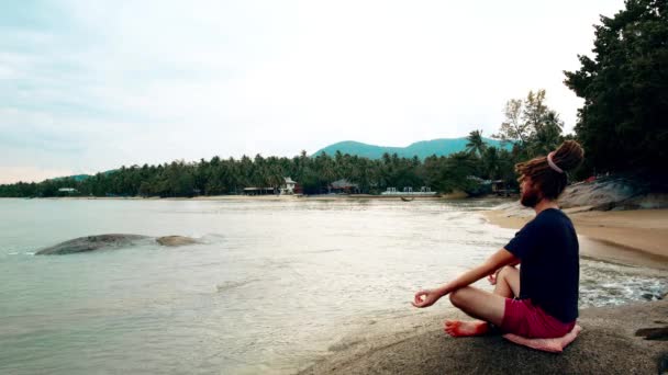 Мужчина медитирует, сидя на камне у моря. Лотос с дредами — стоковое видео