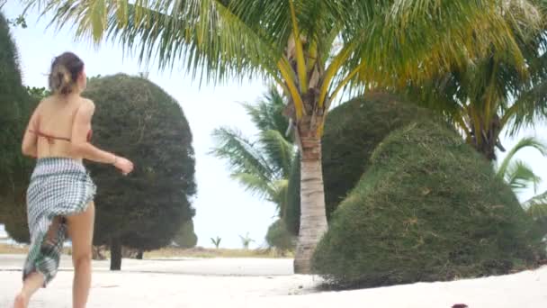 Kobieta na wakacjach biegnie wzdłuż plaży z białym piaskiem i palmami. — Wideo stockowe