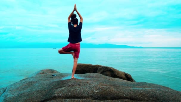 Een man beoefent yoga op grote stenen bij de zee — Stockvideo
