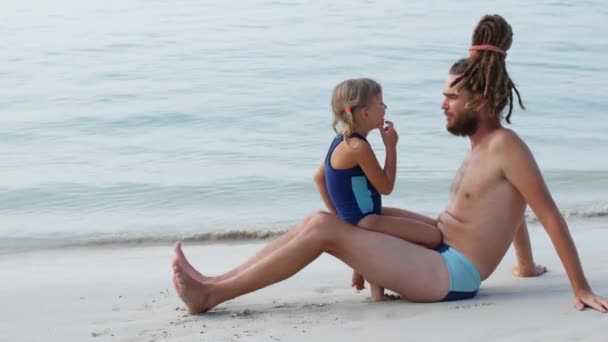 Daughter and father are sitting on the sand by the sea. Girl shows that dad has something stuck in his teeth — 비디오