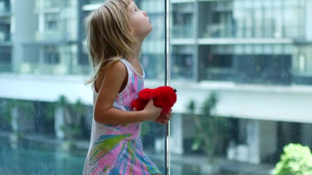Kleines blondes Mädchen steht und wendet sich dem transparenten Glas zu — Stockvideo