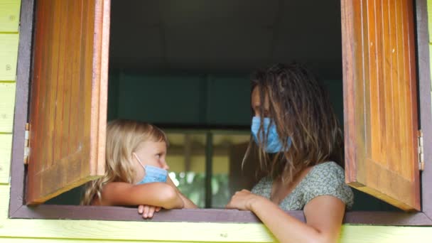 Mamá e hija con máscaras protectoras en la ventana. Pandemia, cuarentena, virus — Vídeos de Stock