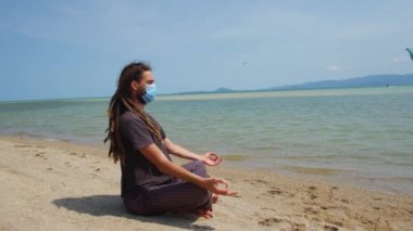 Maskeli bir genç adam sahilde meditasyon yapıyor. Coronavirüs salgını