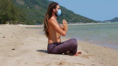 Maskeli bir genç adam sahilde meditasyon yapıyor. Coronavirüs salgını
