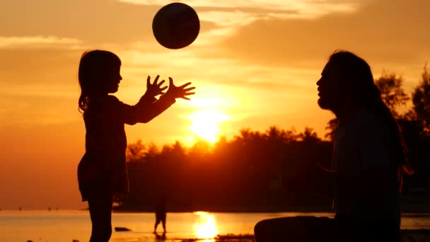 Papá e hija juegan a la pelota en la orilla del mar. Siluetas al atardecer — Vídeo de stock