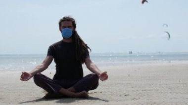 Maskeli bir genç adam sahilde meditasyon yapıyor. Coronavirüs salgını