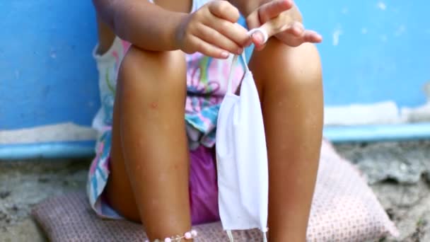 A little girl sits on the ground and twirls a medical mask in her hands. Coronavirus 2010 — Stock Video