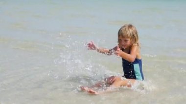 6 yaşında bir kız sığ sularda denizde oturur ve su sıçratır.