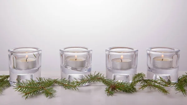Four Advent candles in glass candlesticks on a white background, Christmas spruce branches. The imminent background of Advent and Christmas