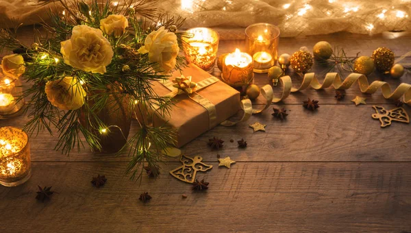 Christmas decoration. Gift, candles, lights, golden balls on a wooden rustic table. Composition of pine branches and English roses in a vase. — Stock Photo, Image