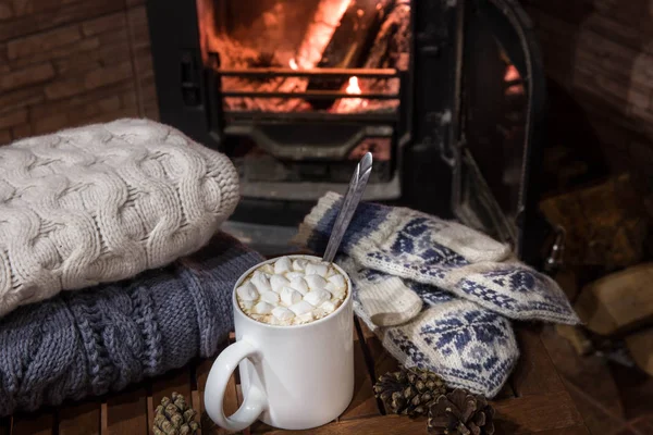 Stack of cozy knitted sweaters, handmade mittens and cup of coffee with marsh mallows on old wooden table, near burning fireplace — Stock Photo, Image