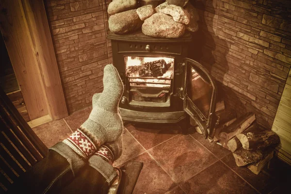 Ноги в шерстяных носках у рождественского камина. Мужчина расслабляется теплым огнем с чашкой горячего напитка и согревает ее ноги шерстяными носками. Зимние и рождественские праздники . — стоковое фото