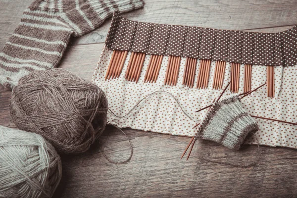 The process of knitting sock with circular bamboo knitting needles, glasses, woolen yarn of natural color, glasses on a wooden rustic table.