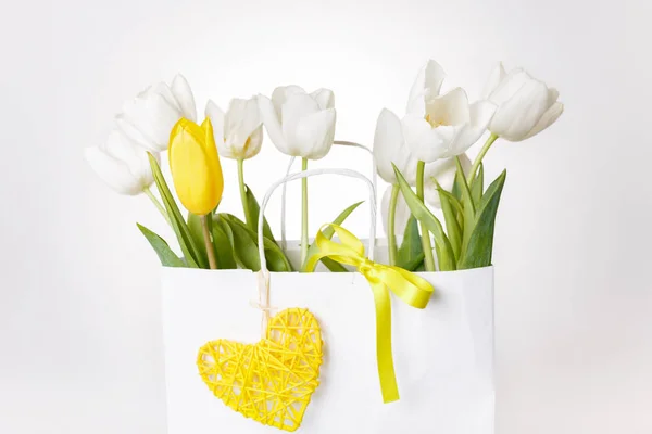 Festive white, yellow tulips composition, handmade heart, ribbon on white background. Bouquet of spring flowers in white paper bag. — Stock Photo, Image