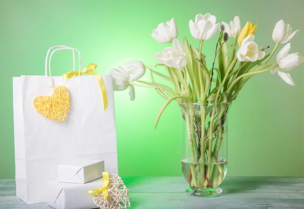 Shopping bag, cuore giallo e nastro, regalo e tulipani bouquet bianchi in vaso su sfondo verde . — Foto Stock