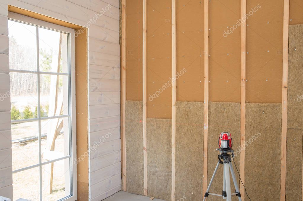 Construction worker thermally insulating eco-wood frame house with wood fiber plates and heat-isolating natural hemp material. Finishing the walls with a white wooden board, using laser line level