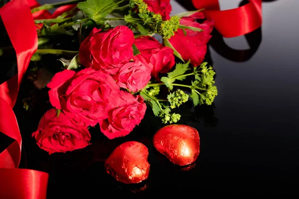 Ramo de rosas rojas y chocolates en forma de corazón, cinta roja sobre un fondo negro — Foto de Stock