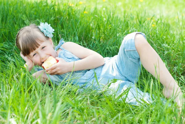 Vacker flicka äter rött äpple på gräsmark. Happy kids concept. — Stockfoto