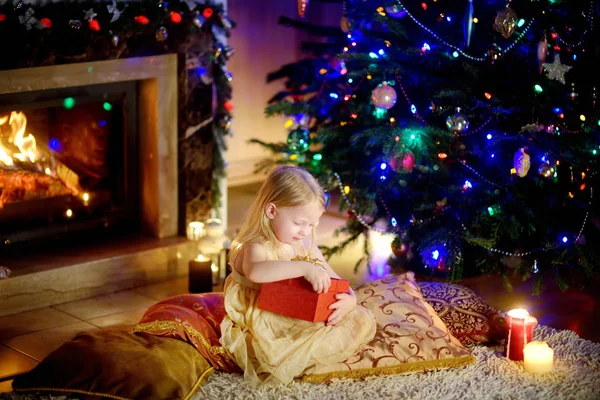 Girl opening Christmas gift by a fireplace — Stock fotografie