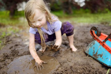 Funny little girl playing in a large wet mud puddle on sunny summer day. Child getting dirty while digging in muddy soil. Messy games outdoors.
