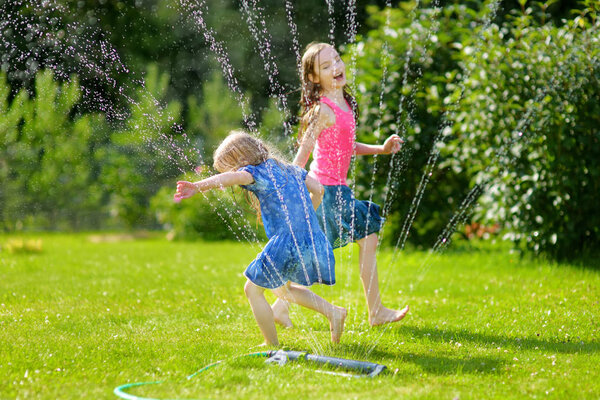 Adorable little girls playing with a sprinkler in a backyard on sunny summer day. Cute children having fun with water outdoors. Funny summer games for kids.