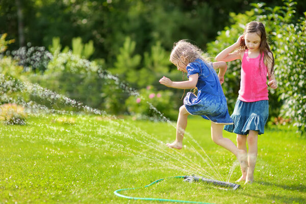 Adorable little girls playing with a sprinkler in a backyard on sunny summer day. Cute children having fun with water outdoors. Funny summer games for kids.